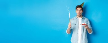 Mutlu genç adam doğum gününü parti şapkasıyla kutluyor, doğum günü pastası tutuyor ve gülümsüyor, mavi arka planda dikiliyor..