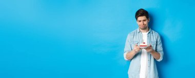 Tatiller ve kutlamalar. Hayal kırıklığına uğramış ve üzgün bir adam doğum günü pastasına bakıyor, üzgün, mavi arka planda duruyor..