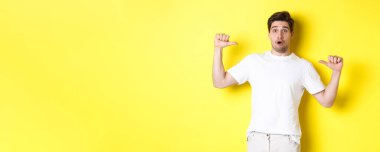 Şaşkın yakışıklı adam kendini işaret ediyor, şaşkın şaşkın bakıyor, sarı arka planda duruyor. Boşluğu kopyala