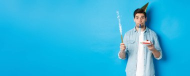 Komik adam doğum gününü kutluyor, elinde doğum günü pastası, havai fişek ve parti şapkası var, mavi arka planda duruyor..