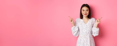 Güzel Asyalı kadın alışverişte seçim yapıyor, parmaklarını yana doğru gösteriyor ve değişkenleri gösteriyor, kameraya gülümsüyor, pembe arka planda duruyor..