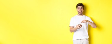 Tanıtım yapmak için akıllı telefonu işaret eden mutlu adam sarı arka planda duran internet teklifini kontrol ediyor. Boşluğu kopyala