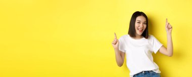 Mutlu Asyalı kadın dans ediyor ve eğleniyor, sarı arka planda beyaz tişörtle poz veriyor..