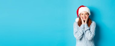 Kış tatili ve Noel arifesi konsepti. Noel Baba şapkalı neşeli kızıl kız, yeni yılı kutluyor, kızarıyor ve gülümsüyor, mavi arka planda duruyor..