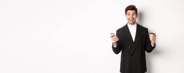 Takım elbiseli mutlu bir adamın portresi, elinde bir fincan kahve ve akıllı telefon, uygulama hedefine ulaşıyor, beyaz arka planda duruyor..