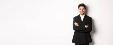 Yakışıklı ve kendine güvenen sakallı bir adam, elleri göğsünde çapraz duran, mutlu bir şekilde gülümseyen, partiye ya da iş etkinliğine katılan, beyaz arka planda duran.