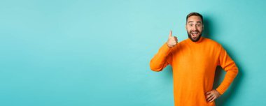 İyi iş çıkardın. Yakışıklı sakallı adam baş parmağını gösterip, iyi işi övüyor, mükemmel bir ürün öneriyor, onaylıyor ve onaylıyor, gülümsüyor, açık mavi arka planda duruyor..