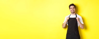 Siyah önlüklü, sarı arka planda duran kendinden emin barista. Garson gülümsüyor ve mutlu görünüyor..