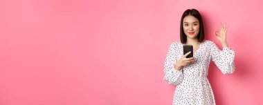 İnternetten alışveriş ve güzellik konsepti. Tatmin olmuş Asyalı bayan müşteri iyi görünüyor, akıllı telefondan internet alımı yapıyor, pembe arka planda duruyor..