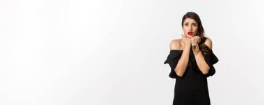 Moda ve güzellik konsepti. Korkmuş ürkek kadın, endişeli ve endişeli görünüyor, siyah elbise içinde beyaz arka planda duruyor..