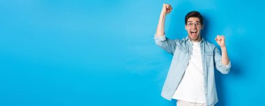 Heyecanlı yakışıklı adam zafer kazanıyor, elini kaldırıyor ve sevinç çığlıkları atıyor, zaferi kutluyor, mavi arka planda duruyor..