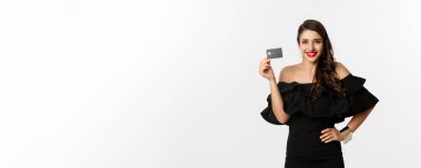 Güzellik ve alışveriş konsepti. Kırmızı dudaklı, siyah elbiseli, mutlu gülümseyen ve kredi kartını gösteren, beyaz arka planda duran zarif bir kadın..