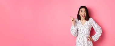 Şık giyinmiş heyecanlı Asyalı kadın, nefes nefese, sol üst köşeyi işaret ediyor, ilginç bir tanıtım teklifi sunuyor, pembe arka planda duruyor..
