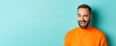 Yakışıklı, kendine güvenen bir adam kameraya göz kırpıyor, küstah gülümsüyor, mavi arka planda turuncu kazak giyiyor..