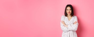 Güzel Asyalı kadın model elbise giymiş, kolları göğsünde ve kameraya gülümsüyor, pembe arka planda duruyor..