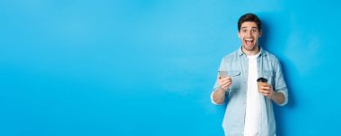 Mutlu genç adam kahve içiyor ve cep telefonu kullanıyor, heyecanlı görünüyor, mavi arka planda duruyor..
