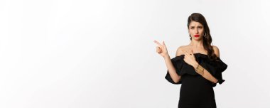 Moda ve güzellik. Hayal kırıklığına uğramış cazibeli kadın şikayet ediyor, siyah elbise giyiyor, somurtuyor ve kötü teklifi işaret ediyor, beyaz arka plan.