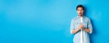 Şaşkın, sıradan giyinmiş, afallamış, akıllı telefon tutan, mavi arka planda duran bir adam..