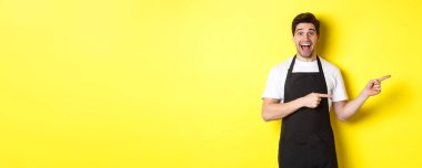 Siyah önlüklü garson sağı işaret ediyor. Reklam veriyor ve heyecanla gülümsüyor. Sarı arka planda duruyor..