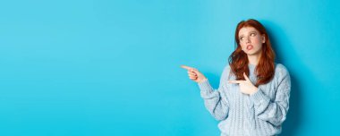 Sinirli, genç, kızıl saçlı kız gözlerini yuvarlıyor, parmaklarını sıkıcı ya da topal bir şeye doğrultuyor, mavi arka planda tedirgin bir şekilde duruyor..