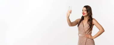 Makyajlı ve saç stili olan güzel bir kadın. Cazibeli bir elbise giyiyor. Akıllı telefondan selfie çekiyor..