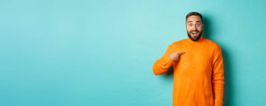 Heyecanlı adam kendini işaret ediyor, şaşkın ve mutlu görünüyor, seçiliyor, açık mavi arka planda duruyor..