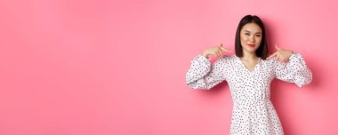 Güzel Asyalı kadın kendinden emin görünüyor, kendini gösteriyor ve gülümsüyor, göğsünde logo var, pembe arka planda duruyor..