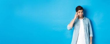 Genç düşünceli bir adam kafasını işaret ediyor, düşünmek istiyor, ipucu veriyor, mavi arka planda günlük kıyafetlerle dikiliyor..