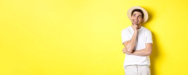 Düşünceli genç adam turist görüntüsü veriyor, sol üst köşeye bakıyor ve gülümsüyor, düşünüyor ve sarı arka planın üzerinde duruyor..