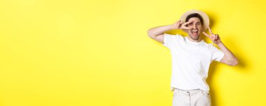 Turizm ve tatil kavramı. Mutlu turist barış işaretleriyle poz veriyor, heyecanlı gülümsüyor, sarı arka planda duruyor..