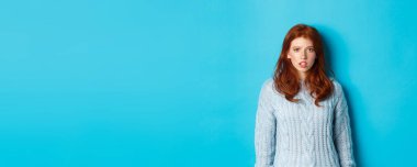 Süveter giymiş kafası karışık bir kızıl kız kameraya bakıyor, kaşlarını kaldırıyor ve şaşkın bir şekilde mavi arka planda duruyor..