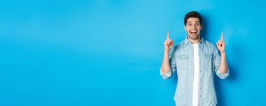 Mutlu 25 'li yaşlarda sakallı, parmaklarını kaldırıp gülümseyen, reklamını yapan, mavi arka planda duran bir adamın portresi..