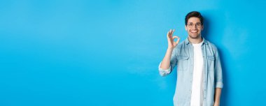 Gözlüklü ve günlük kıyafetler giyen çekici bir genç. Onaylama konusunda iyi bir işaret veriyor. Mavi arka plana karşı duran bir şey..