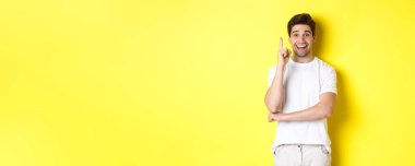 Çekici bir adamın fikri olduğunu hayal etmek, parmağını kaldırıp plan önermek, heyecanlı gülümsemek, sarı arka planda dikilmek..