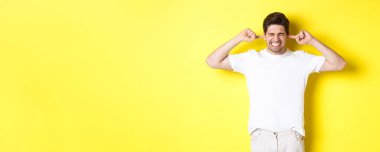 Sinirli adam suratını buruşturup kulaklarını kapatıyor, gürültüden şikayet ediyor, sarı arka planda duruyor. Boşluğu kopyala