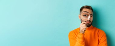 Komik adam büyüteçten bakıyor, bir şeyi arıyor ya da araştırıyor turkuaz arka planda turuncu süveter giyiyor..