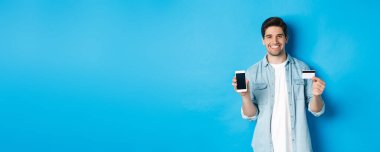 Akıllı telefon ekranı ve kredi kartı gösteren gülümseyen genç adam, online alışveriş veya bankacılık kavramı.