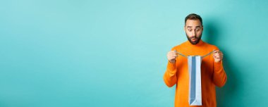 Şaşırmış bir adam alışveriş çantasını açtı ve şaşkın görünüyordu, tatilde hediye alıyordu, turkuaz arka planda duruyordu..