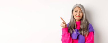 Pembe kazaklı hoşnutsuz Asyalı büyükanne, somurtuyor ve somurtuyor şüpheci, sol üst köşesini işaret ediyor, beyaz arka planda duruyor..
