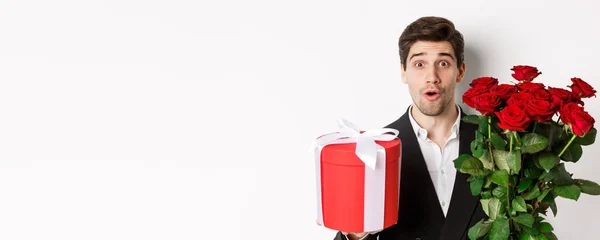 stock image Close-up of attractive man in suit looking surprised, holding gift box and bouquet of roses, giving present for holiday, standing against white background.
