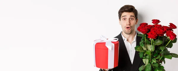 Stock image Close-up of attractive man in suit looking surprised, holding gift box and bouquet of roses, giving present for holiday, standing against white background.