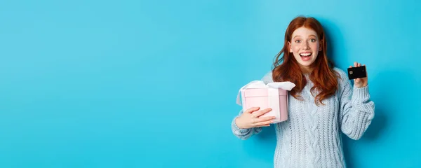 Winter holidays promo offer concept. Cheerful redhead woman holding Christmas gift and credit card, staring at camera amazed, standing over blue background.