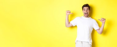 Heyecanlı adam mutlu görünüyor, kendini hayretle işaret ediyor, sarı arka planda duruyor..