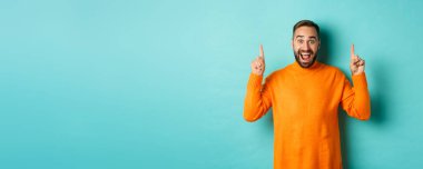 Young happy man showing advertisement, pointing fingers up announcing promo offer, standing over turquoise background.