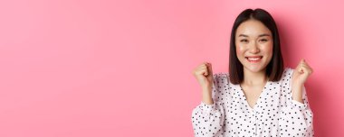 Güzellik ve yaşam tarzı konsepti. Kutlama yapan neşeli Asyalı kadının yakın çekimi, yumruk sallaması ve sevinç gülümsemesi, kazanmak ve zafer, pembe arka planda durmak.
