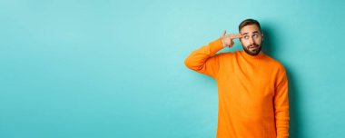 Adam kendini kafasından vuruyor, parmak tabancasını şakağa yaklaştırıyor ve gözleri yuvarlanıyor, açık mavi arka plana karşı koymaktan rahatsız oluyor..