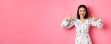Güzel Koreli kadın logonuzu işaret ediyor ve gülümsüyor. Romantik pembe arka planda giyinmiş duruyor. Boşluğu kopyala