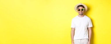 Turizm ve tatil kavramı. Rahatlamış gülümseyen adam akşam yemeğinin tadını çıkarıyor, güneş gözlüğü ve hasır şapka takıyor, sarı arka plan.