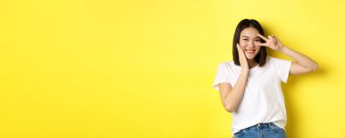 Beyaz tişörtlü sevimli Asyalı kız. Elleri yanağında poz veriyor. Gözlerinde barış işareti, sarı arka planda duruyor..
