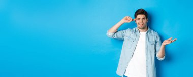 Kafası karışmış bir adam aptal olduğu için insanları azarlıyor, kafasını işaret ediyor ve omuz silkiyor, şaşkın görünüyor, onaylanmamış eylemler yapıyor, mavi arka planda duruyor..